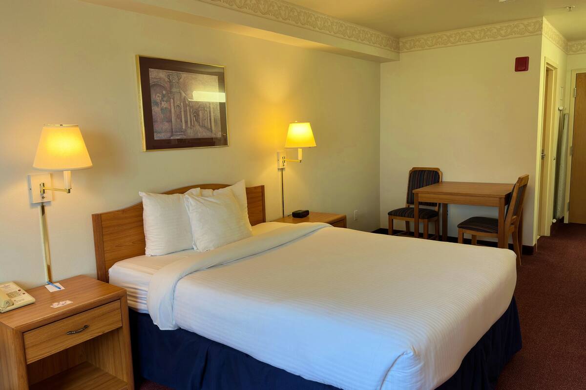 A hotel room with a large bed, two bedside lamps, a desk with a chair, and a framed picture on the wall.