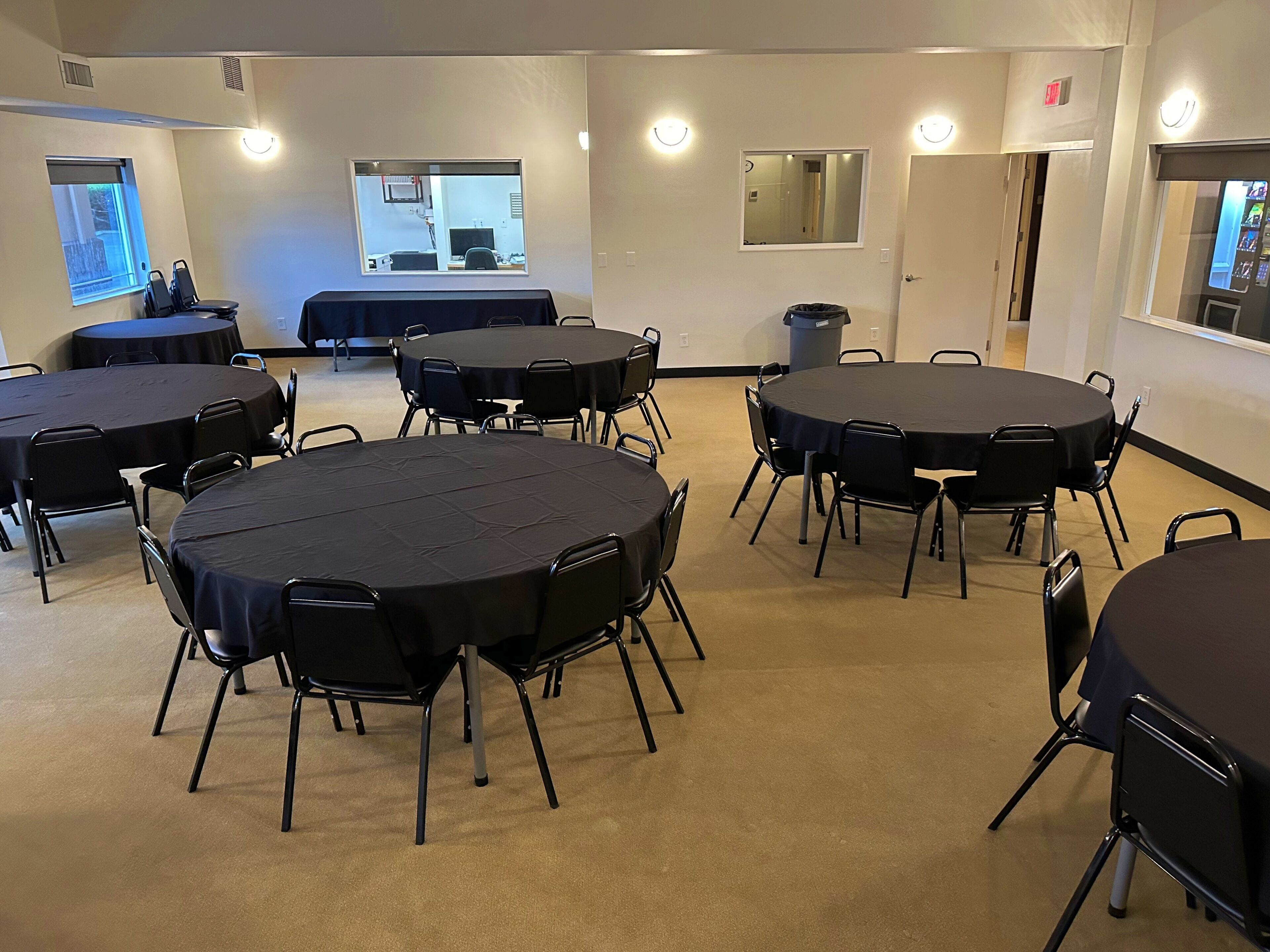 A conference room with multiple round tables and chairs, a trash bin, and a window with a view.