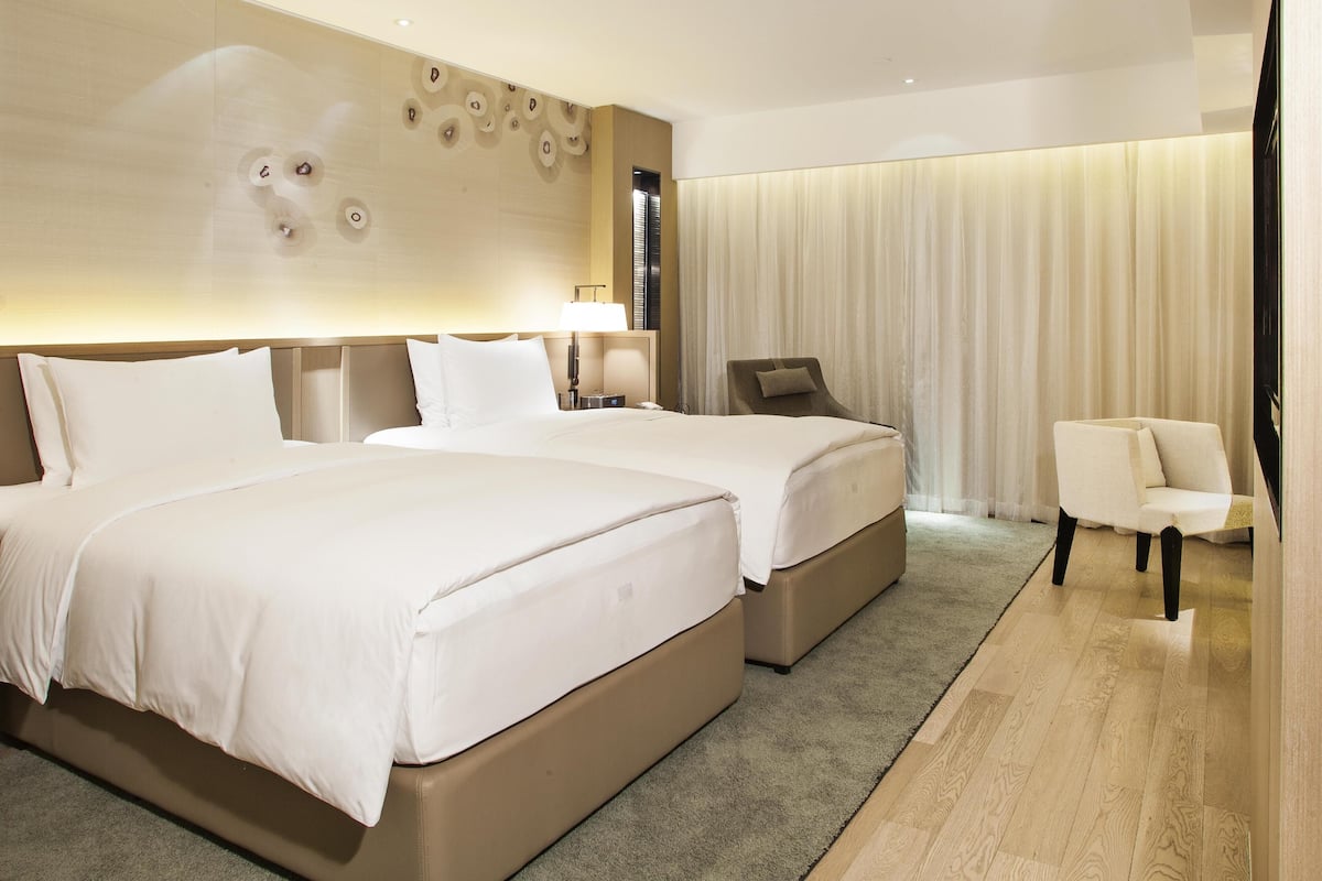 Een hotelkamer met twee bedden, een zithoek met een stoel en een groot raam met gordijnen.