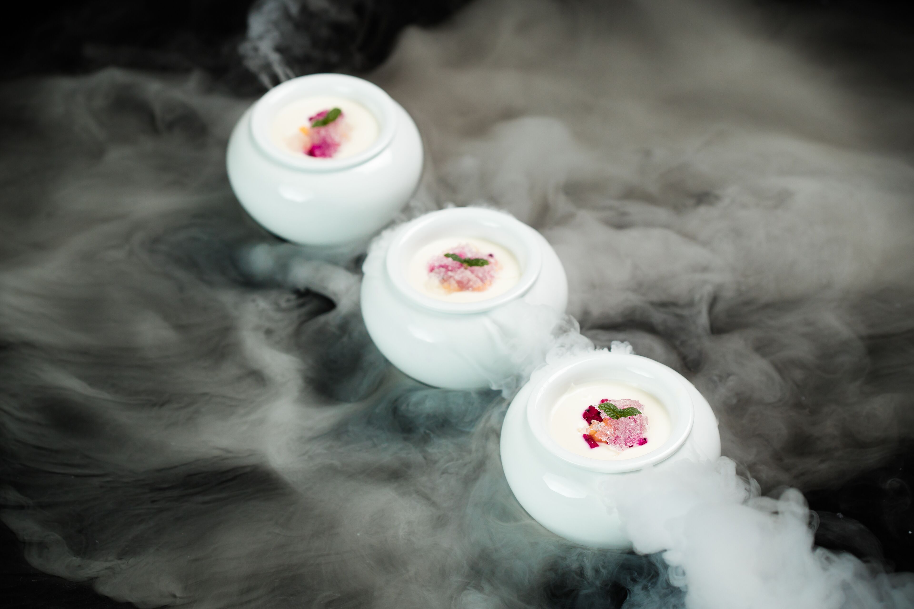Three white teapots with a pink substance and mint leaves on a smoky background.