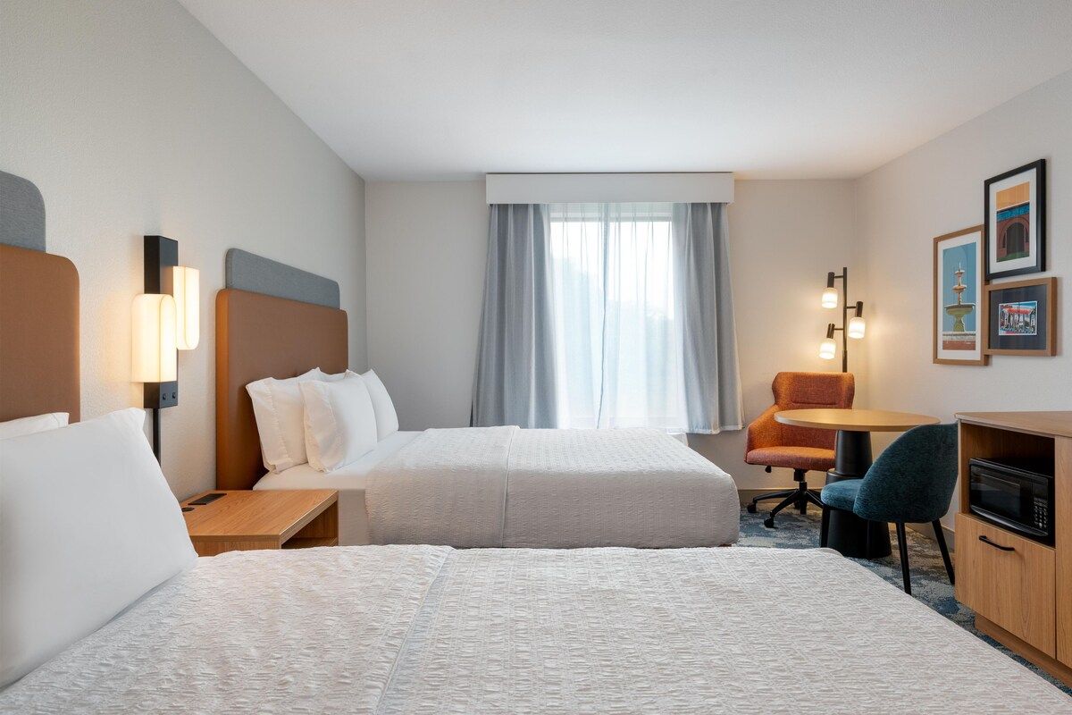 Egy szállodai szoba két ággyal, íróasztallal, székkel, mikrohullámú sütővel és függönnyel ellátott ablakkal.