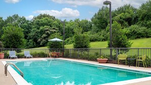 Une piscine rectangulaire avec un escalier en métal, entourée d’une clôture, et un espace extérieur aménagé avec des chaises et des tables.