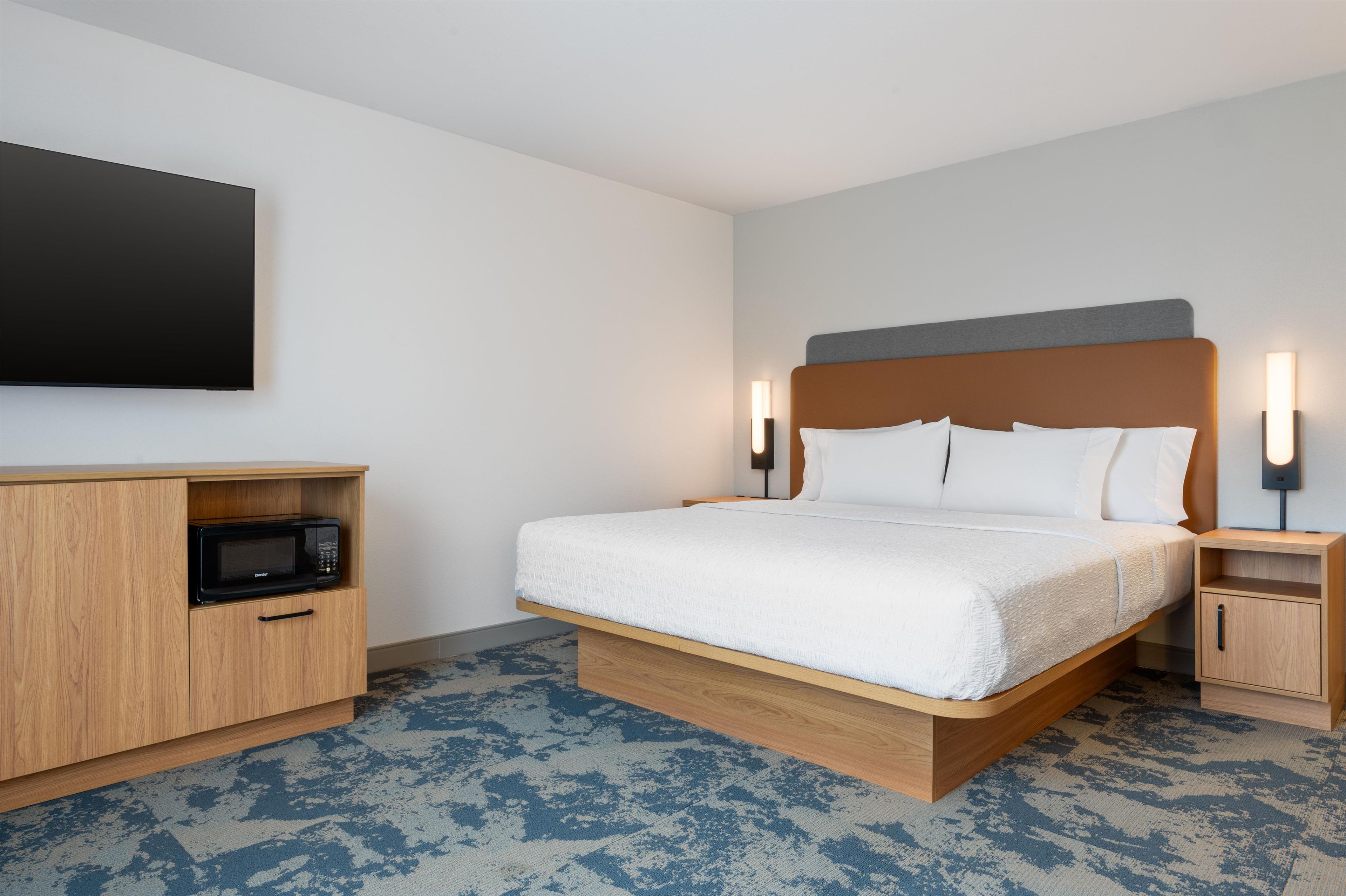 Une chambre d’hôtel comprenant un lit, un téléviseur à écran plat fixé au mur, un four à micro-ondes dans un meuble en bois, et deux tables de chevet avec des lampes.
