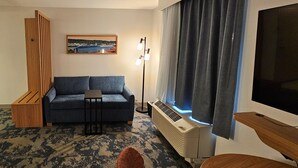 A hotel room with a sofa, a small table, a standing lamp, a large window with curtains, a flat-screen TV, and a radiator. - Hampton Inn & Suites Prattville (Prattville)