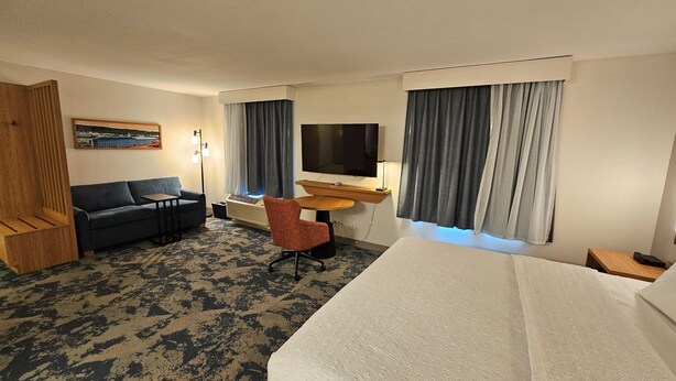 A hotel room with a bed, a sofa, a desk with a flat-screen TV, a chair, and a large window with curtains. - Hampton Inn & Suites Prattville (Prattville)