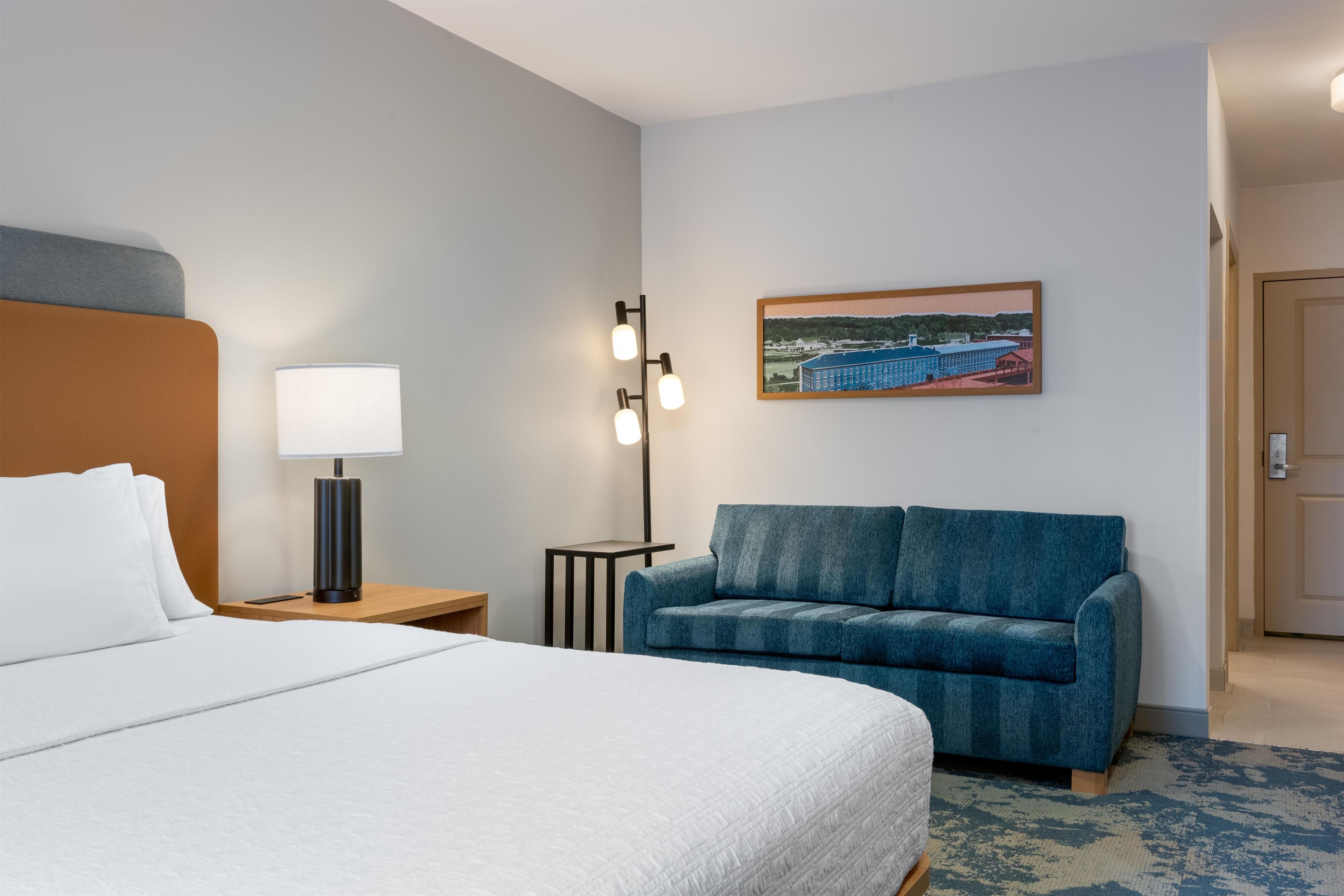 A hotel room with a bed, a desk, a lamp, a small table, a blue sofa, and a painting on the wall.