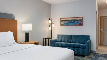 A hotel room with a bed, a desk, a lamp, a small table, a blue sofa, and a painting on the wall.