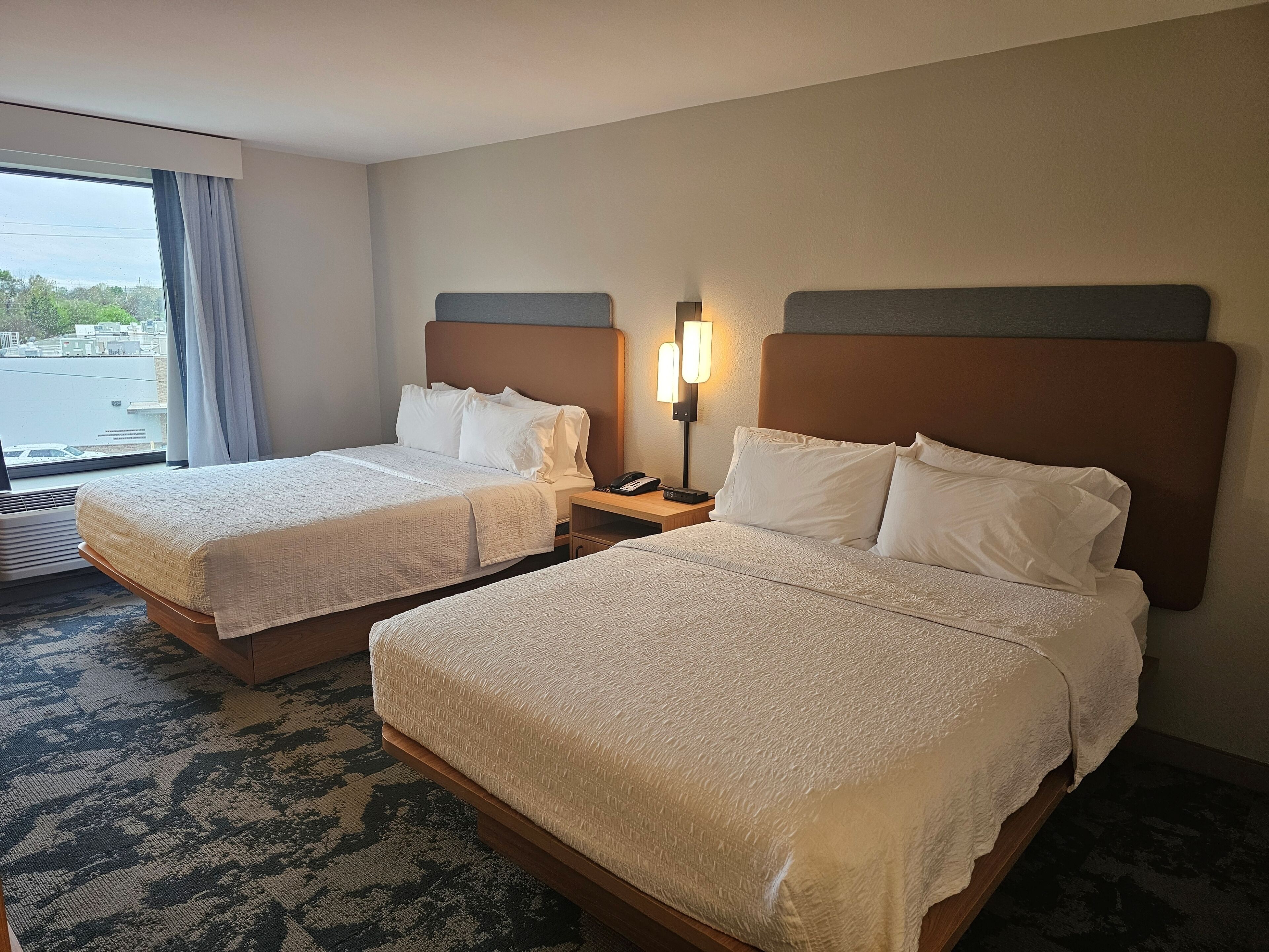 A hotel room with two beds, a nightstand with a lamp, and a view of a body of water through the window.
