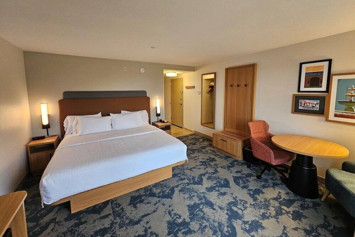 Habitación de hotel con cama, mesitas de noche, una silla, una pequeña mesa, un espejo y cuadros en la pared.