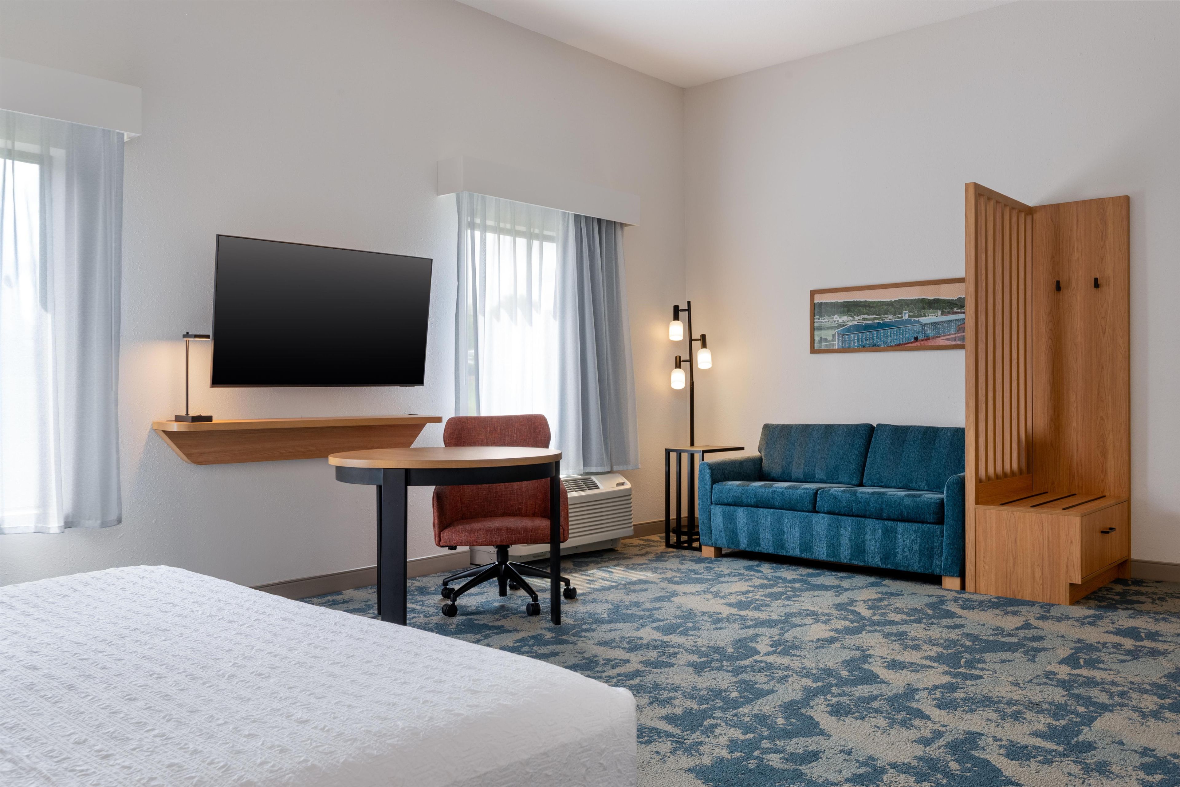 A hotel room with a bed, a desk with a chair, a sofa, a TV, and a painting on the wall.