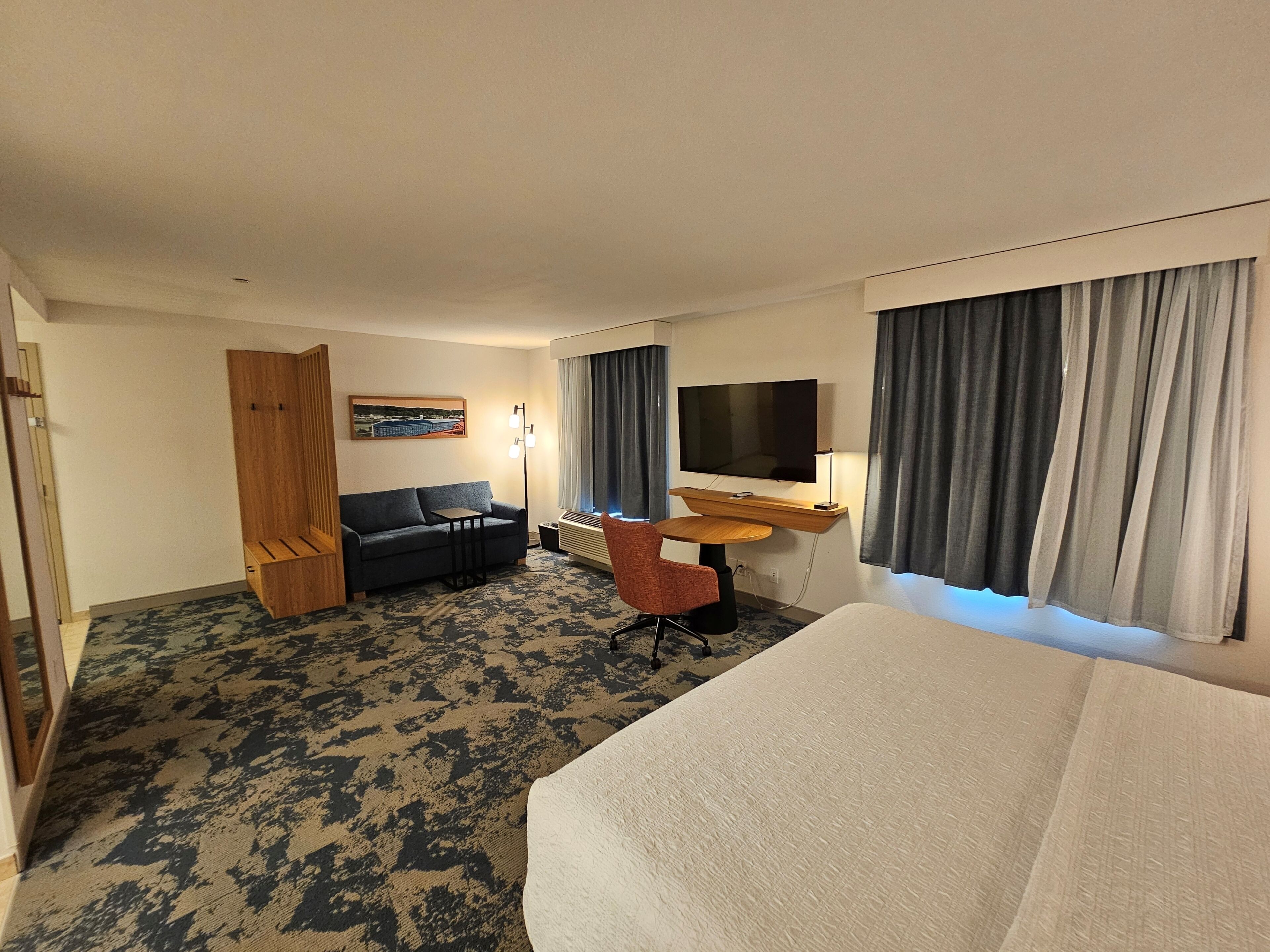 A hotel room with a bed, a sofa, a desk with a TV, a chair, and a painting on the wall.