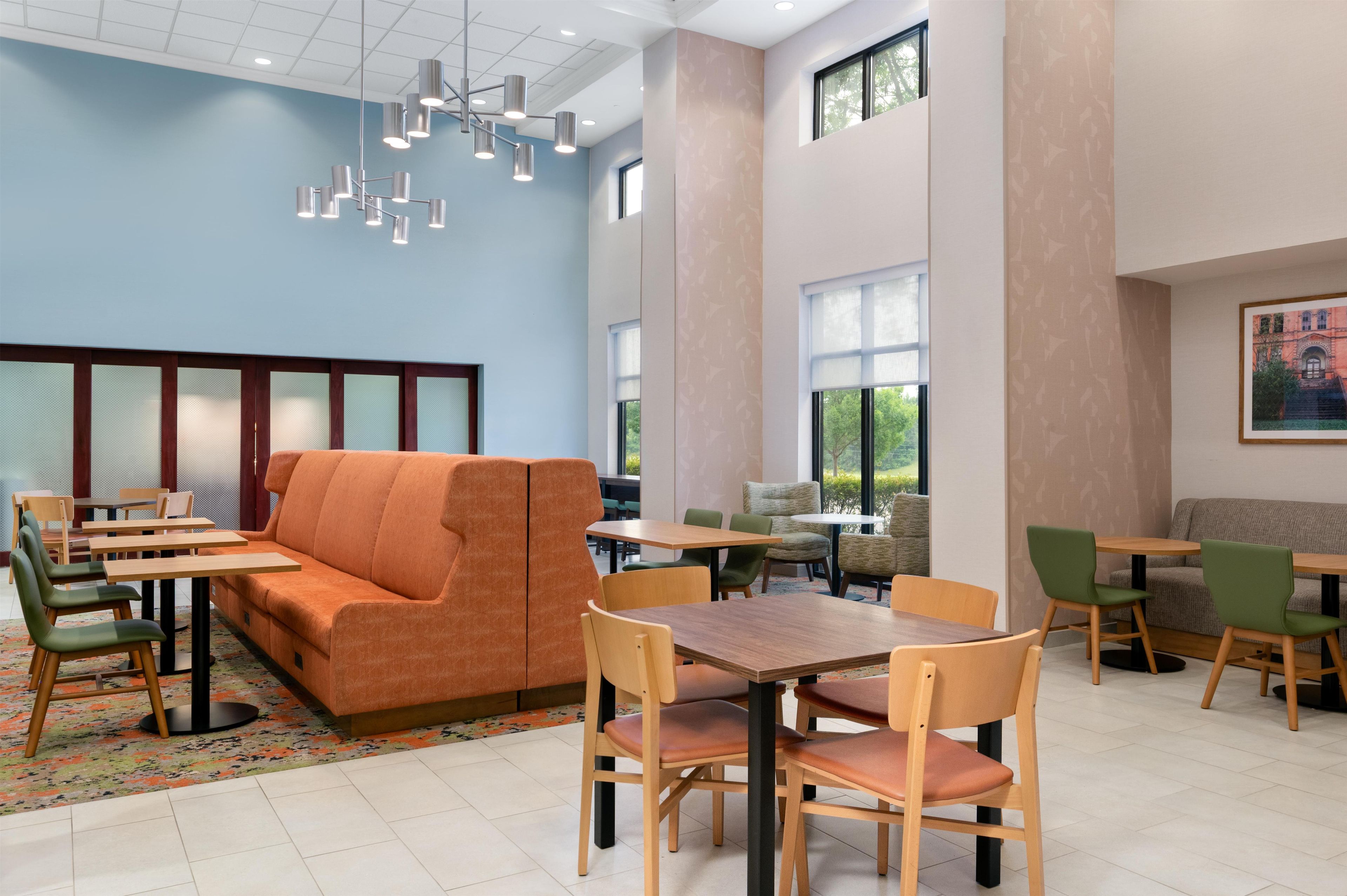 A modern hotel lobby with a sofa, dining tables, chairs, and a large window providing a view of the outdoors.