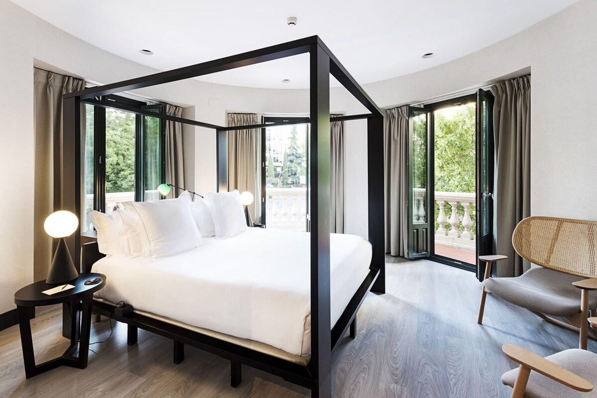 A modern hotel room with a four-poster bed, a sitting area with a chair, a small table, and a balcony with curtains.
