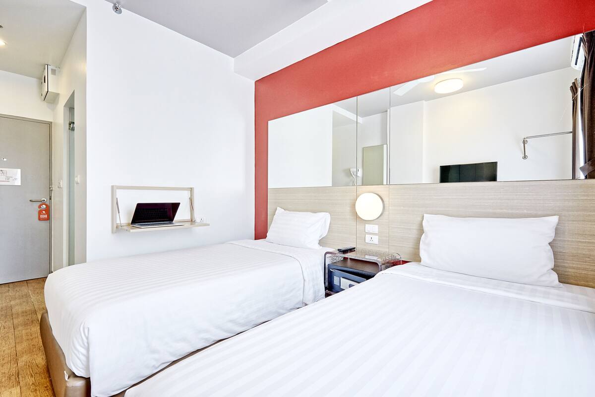 A hotel room with two beds, a laptop on a bedside table, a mirror, and a red accent wall.