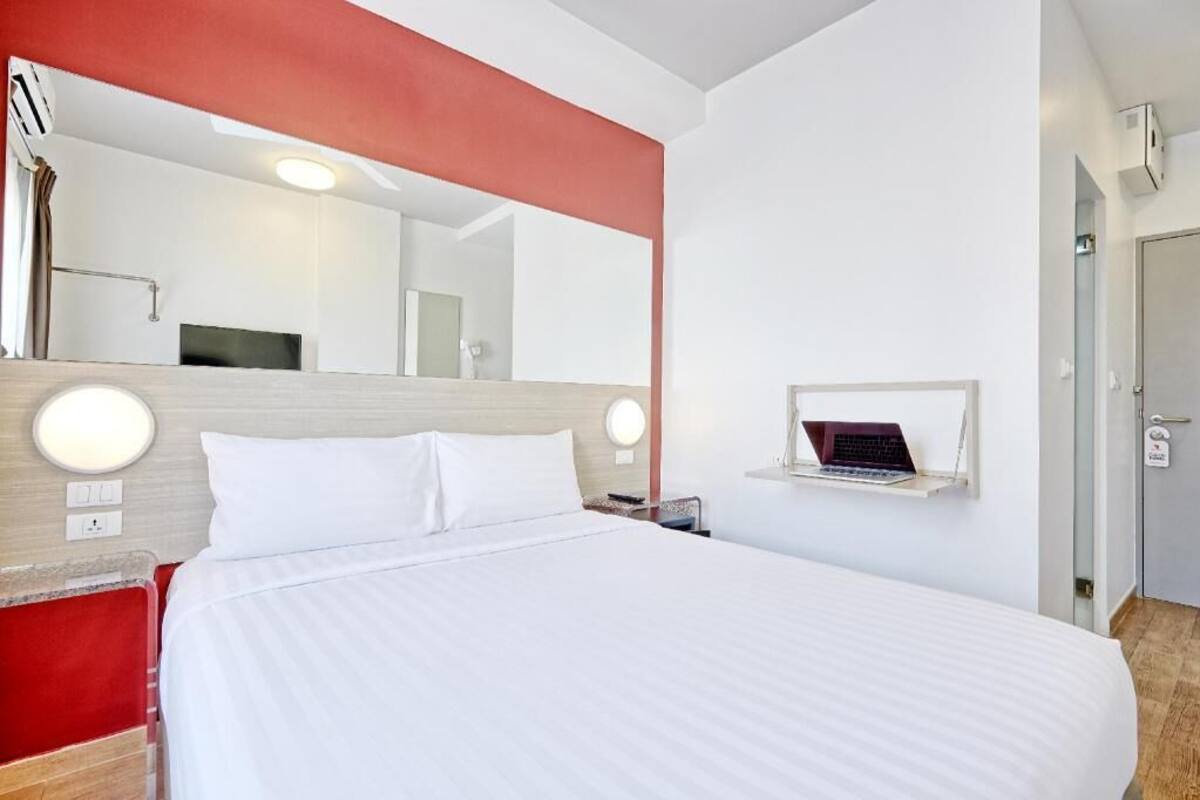 A hotel room with a bed, a desk with a laptop, a mirror, and a red accent wall.