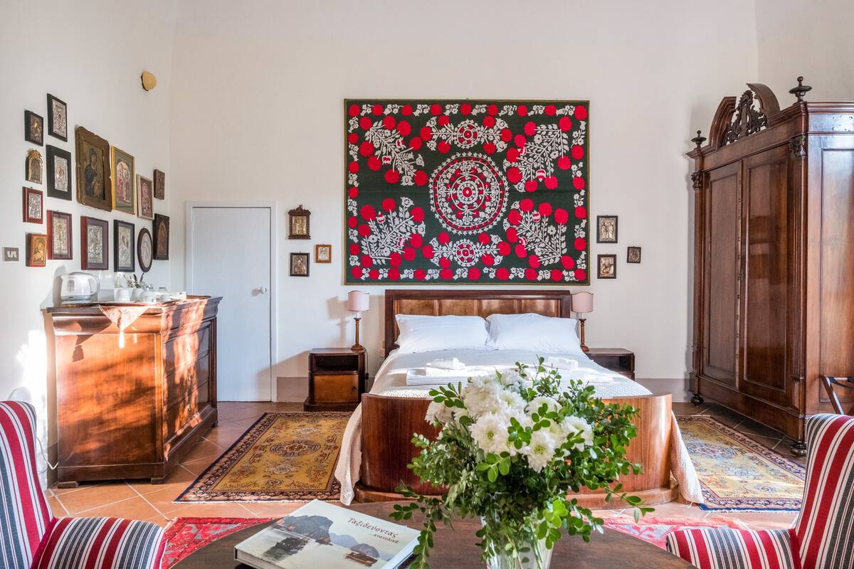 A bedroom with a bed, a dresser, a nightstand, a lamp, a wooden wardrobe, a wall with framed pictures, and a large decorative tapestry.
