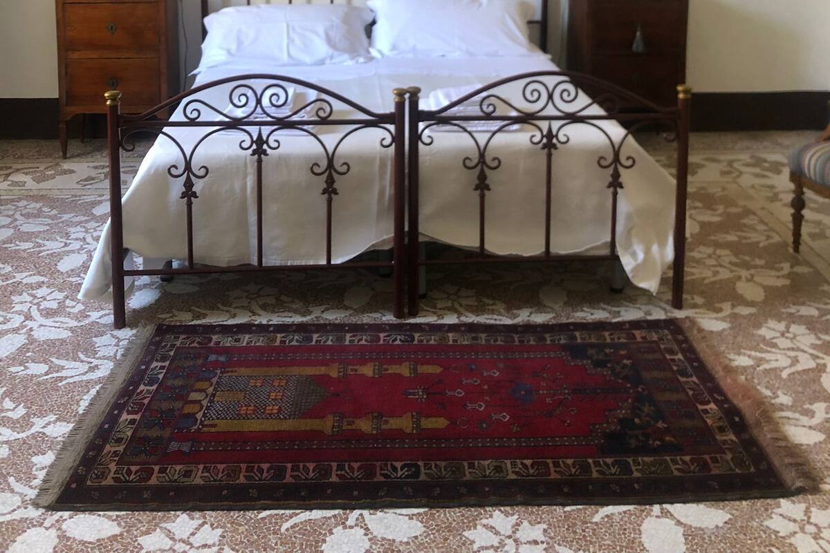 A bedroom with a four-poster bed, a patterned rug, a wall hanging, and a nightstand with a lamp.
