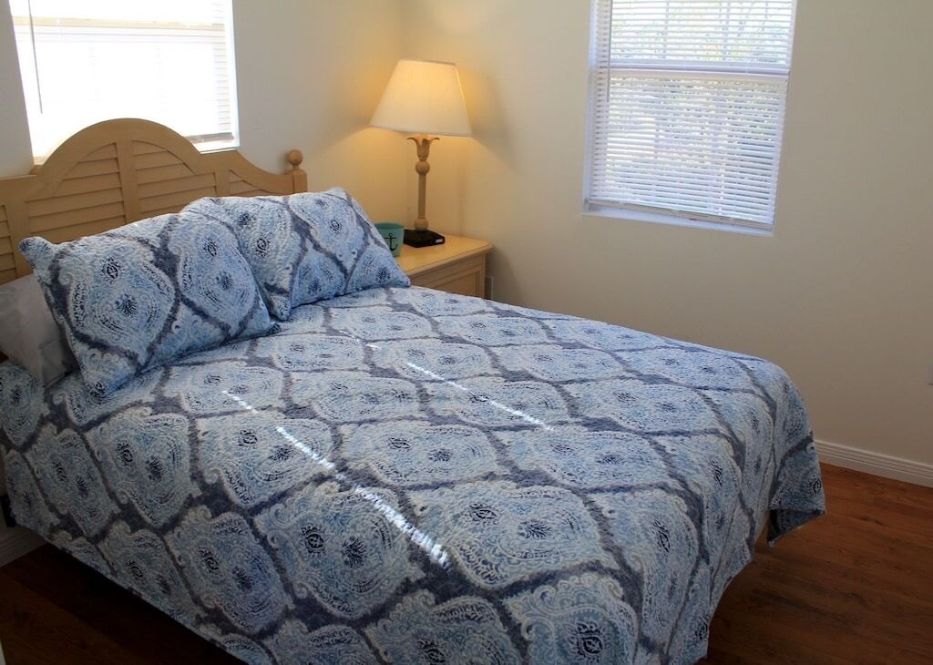 Un dormitorio con cabecera de madera, colcha estampada, lámpara de noche y ventana con persianas.