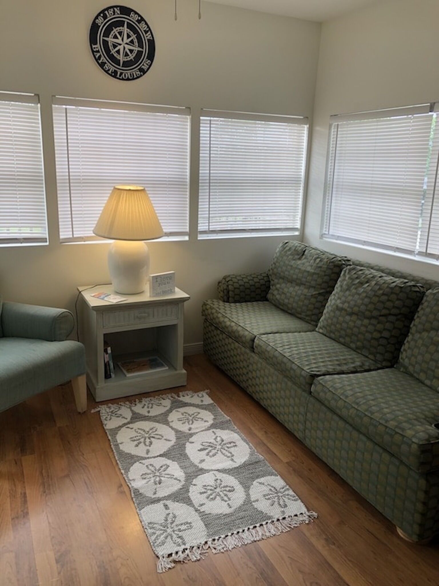 A living room with a green patterned sofa, a small white side table with a lamp, a blue armchair, and a decorative rug.