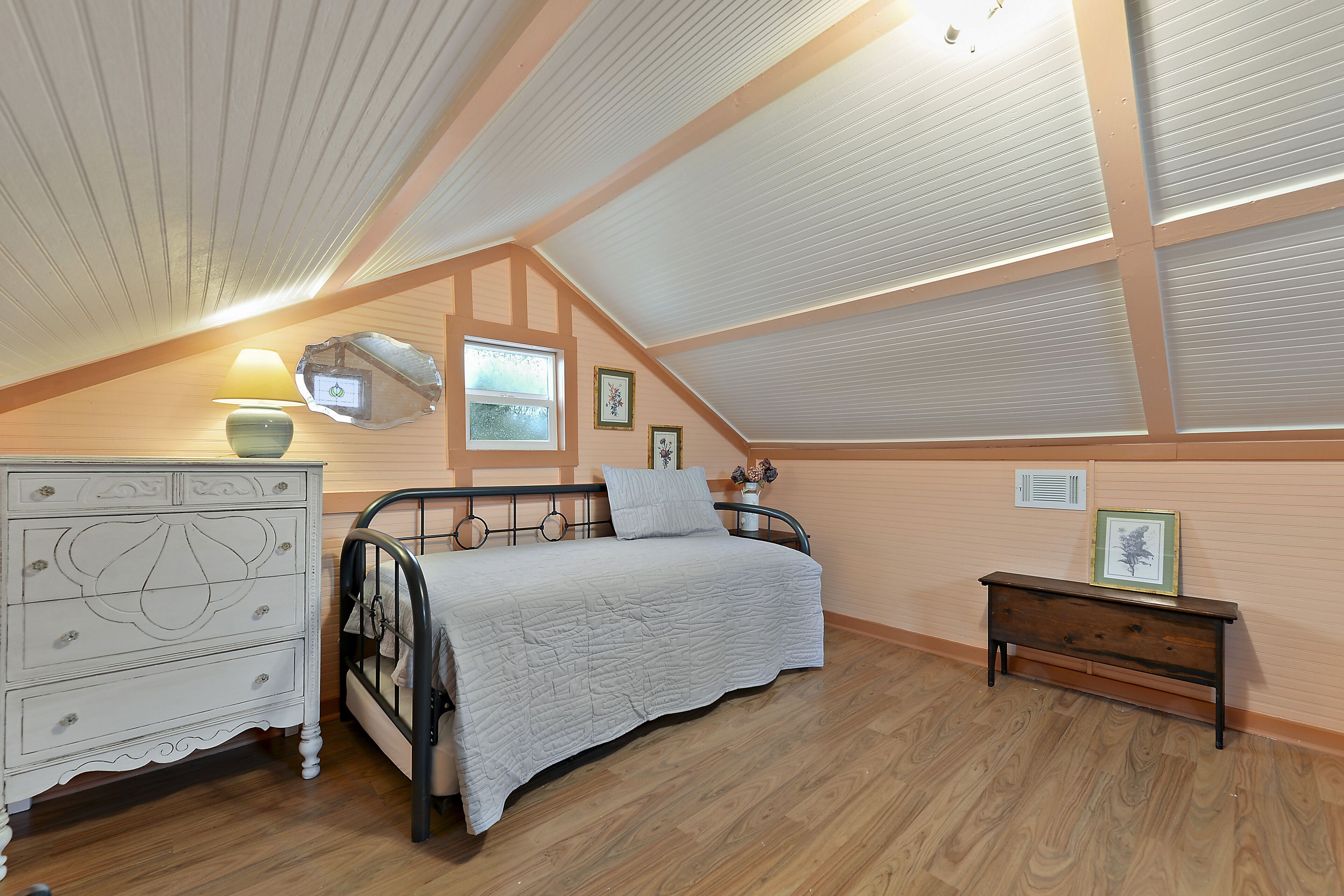 A bedroom with a sloped ceiling, a bed with a white quilt, a dresser, and a small bedside table with a lamp.