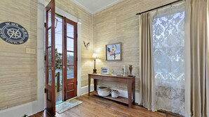 A wooden-floored room with a wooden door, a wall-mounted compass, a framed picture, a table with decorative items, and a patterned curtain. - Carroll House Cottages (Bay St Louis)