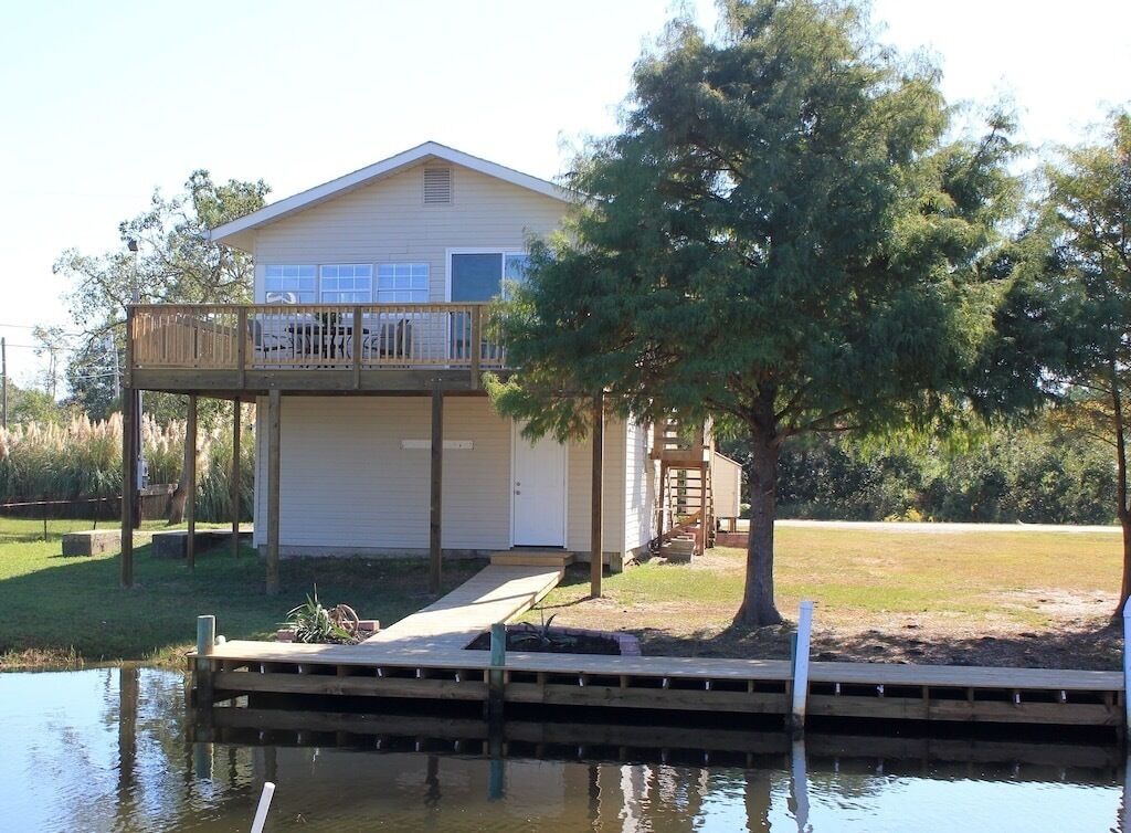 A single-story house with a balcony, a dock extending into a body of water, and a tree in the foreground.