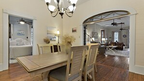 A spacious dining area with a wooden table and chairs, a chandelier, and a view into a bedroom with a bed and curtains. - Carroll House Cottages (Bay St Louis)