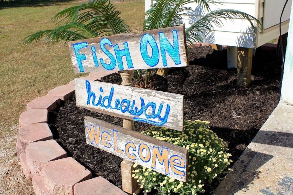 A wooden sign with "FISH ON," "hideaway," and "welcome" written on it, placed in a landscaped area with plants.