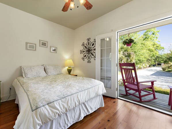 A bedroom with a bed, a nightstand, a lamp, and a door leading to an outdoor area with a rocking chair. - Carroll House Cottages (Bay St Louis)