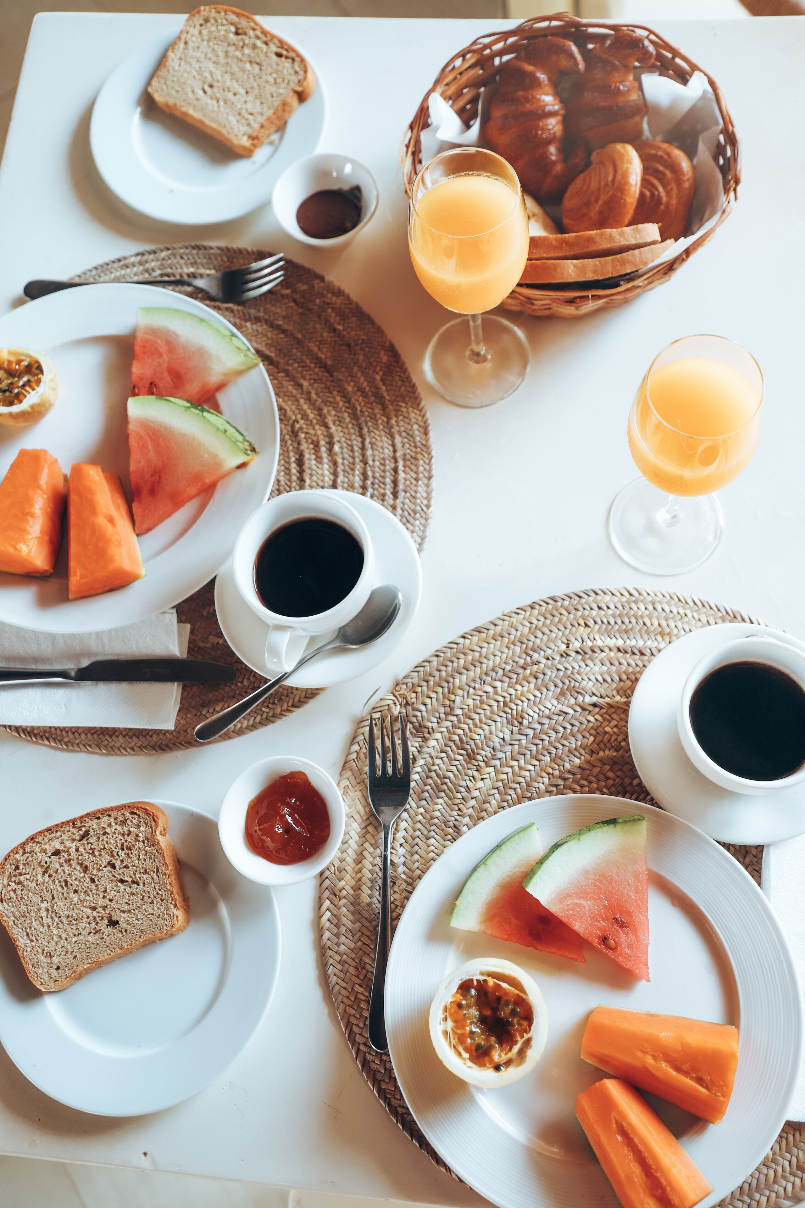 A breakfast table with plates of toast, fruit, and beverages, including a basket of croissants.