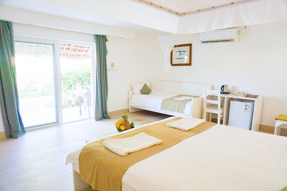 A hotel room with a large bed, a desk, a chair, and a small refrigerator. There is a sliding glass door leading to an outdoor area.
