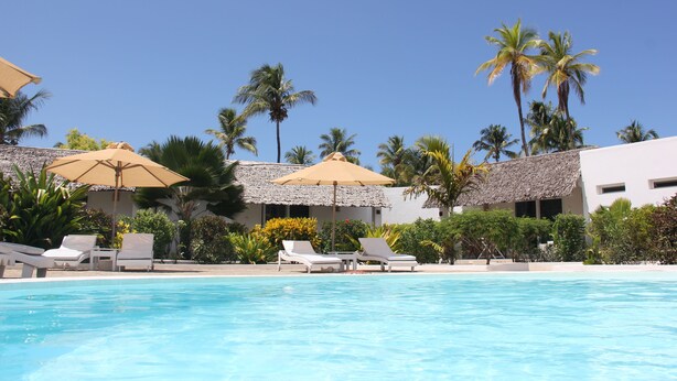 A pool area with lounge chairs and umbrellas, surrounded by palm trees and a building with a thatched roof. - Dhow Inn Boutique Hotel (Paje)