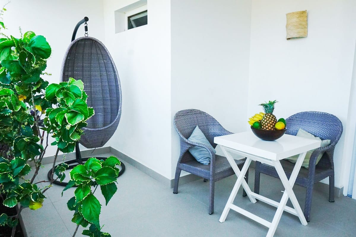 A small balcony with a wicker chair, a white table, and a bowl of fruit.
