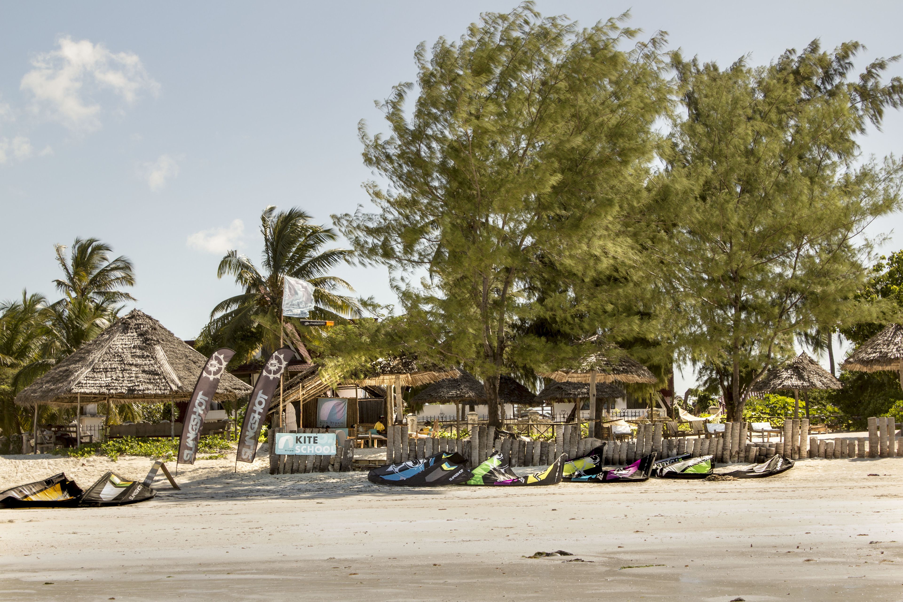 A beach with thatched-roof huts, a sign for 'Kite Choo', and several boats lined up on the sand.