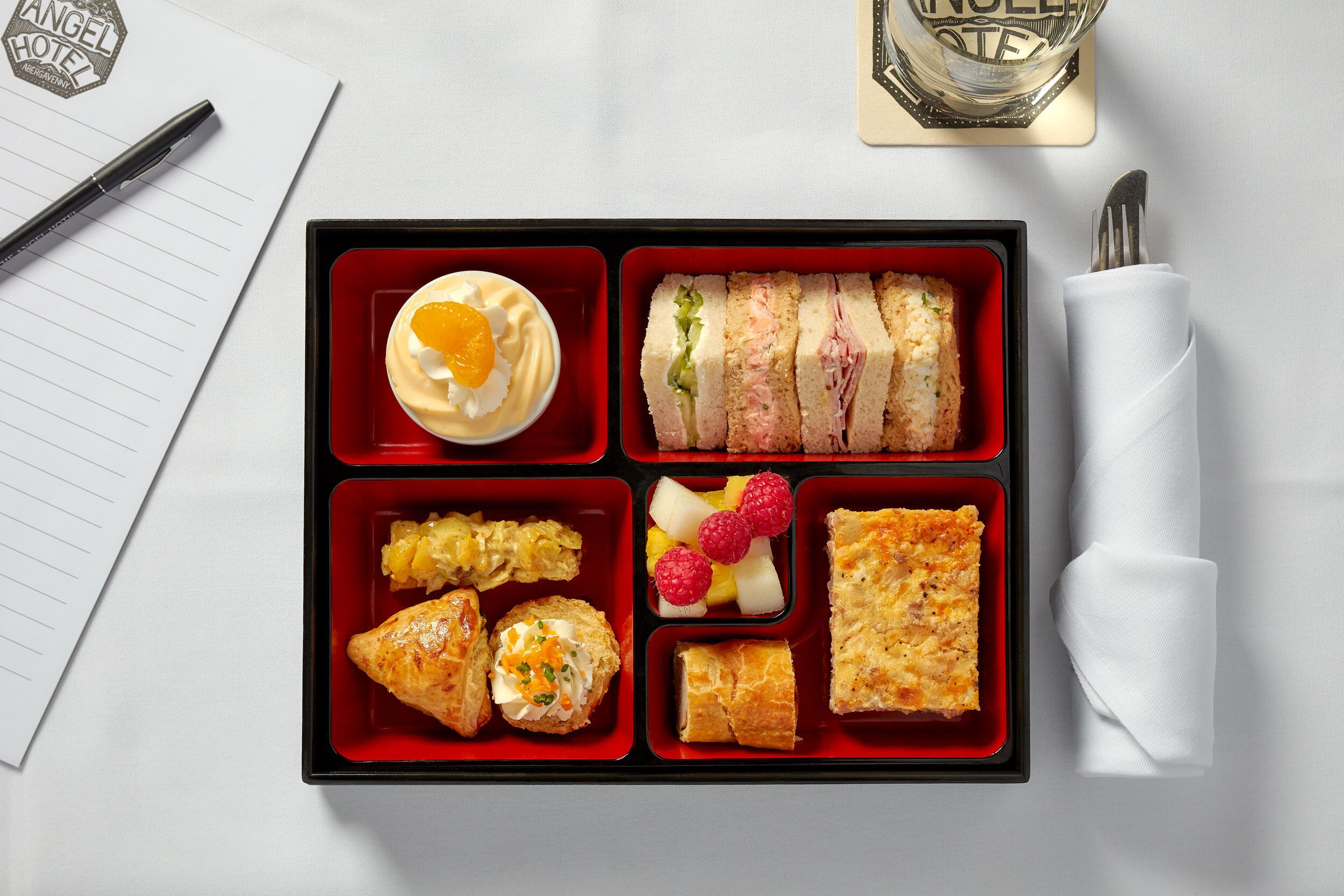 A bento box with various food items, a pen, a napkin, and a glass of water on a table.
