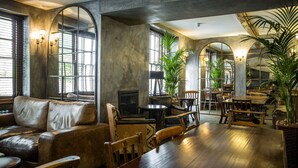 A spacious interior with a wooden dining table, leather sofa, and various chairs. There are large windows with arched frames and several potted plants. - The Angel Hotel Abergavenny (Abergavenny)