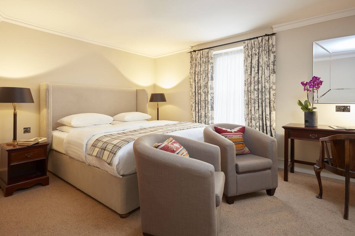 A hotel room with a large bed, two armchairs, a desk, a lamp, a nightstand, and a mirror.
