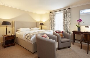 Una habitación de hotel con una cama grande, dos sillones, un escritorio, una lámpara, una mesita de noche y un espejo.