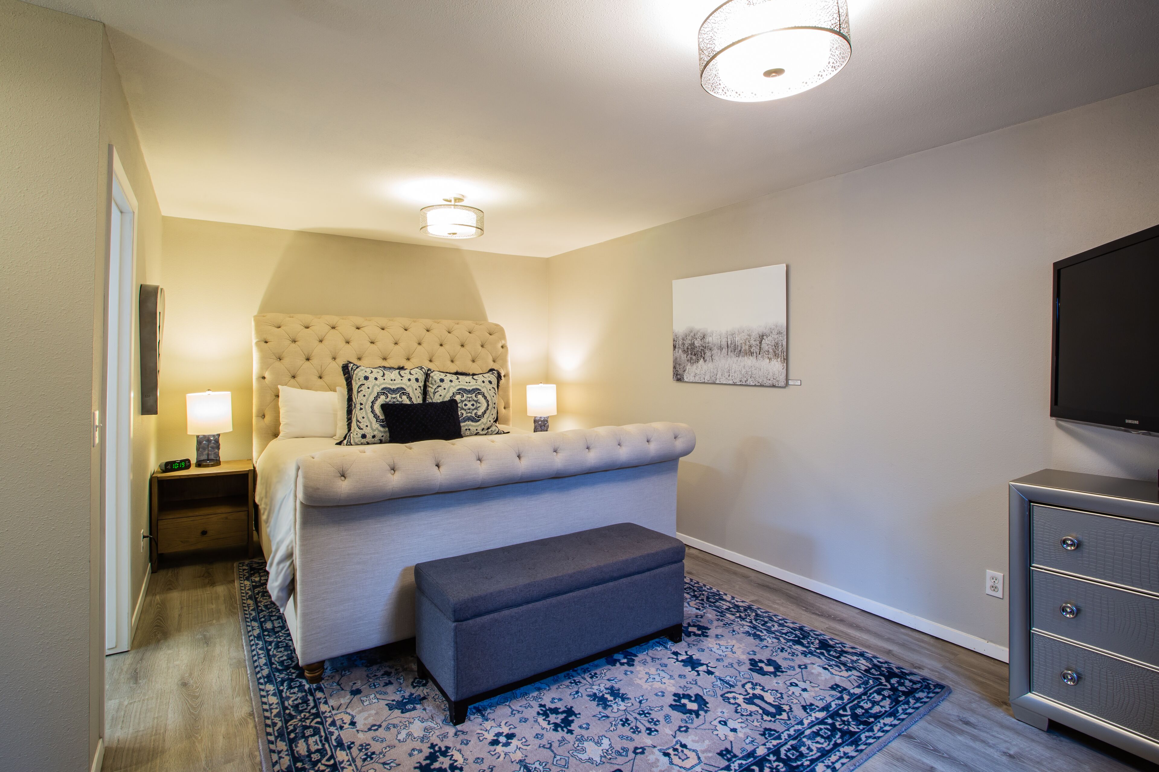 Une chambre à coucher avec un lit doté d’une tête de lit capitonnée, des lampes de chevet, un téléviseur à écran plat, un banc gris et un tapis à motifs.