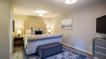 Une chambre à coucher avec un lit doté d’une tête de lit capitonnée, des lampes de chevet, un téléviseur à écran plat, un banc gris et un tapis à motifs.