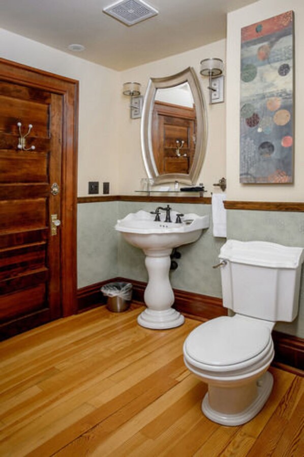 A bathroom with a wooden floor, a pedestal sink, a toilet, and a wooden door.