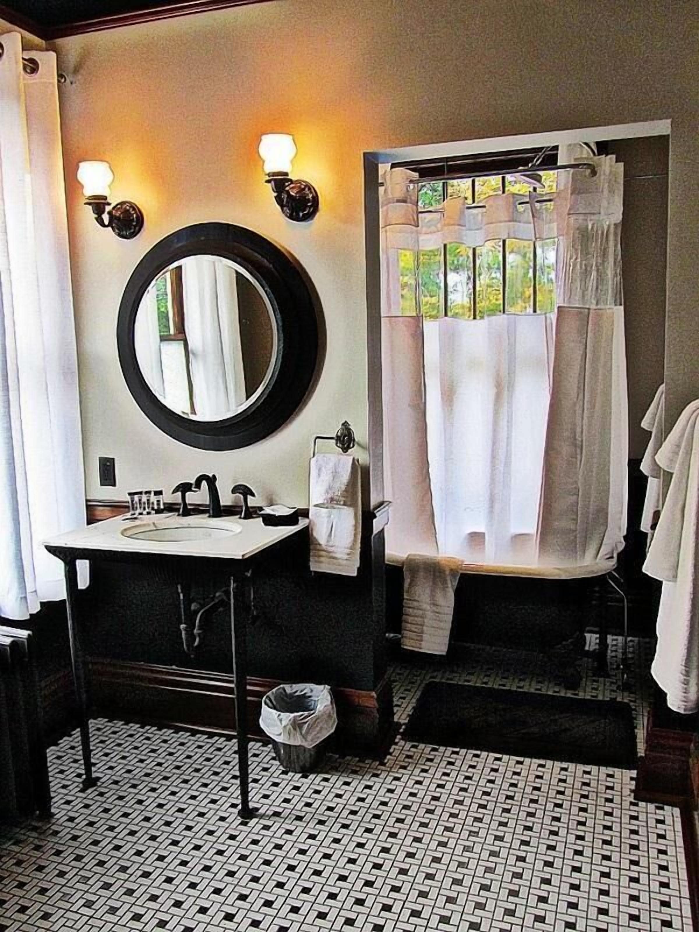 Un baño con un espejo redondo, dos apliques de pared, un lavabo, un perchero para toallas y una cortina de ducha.