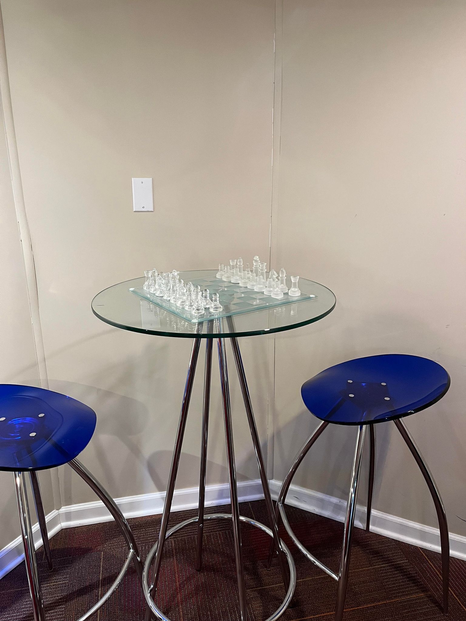 Una mesa con superficie de vidrio y piezas de ajedrez, flanqueada por dos taburetes azules con patas de metal.