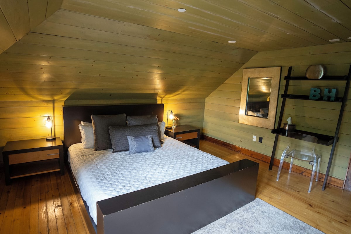 A bedroom with a wooden ceiling, a bed with multiple pillows, bedside lamps, a nightstand, a mirror, and a shelf with letters.