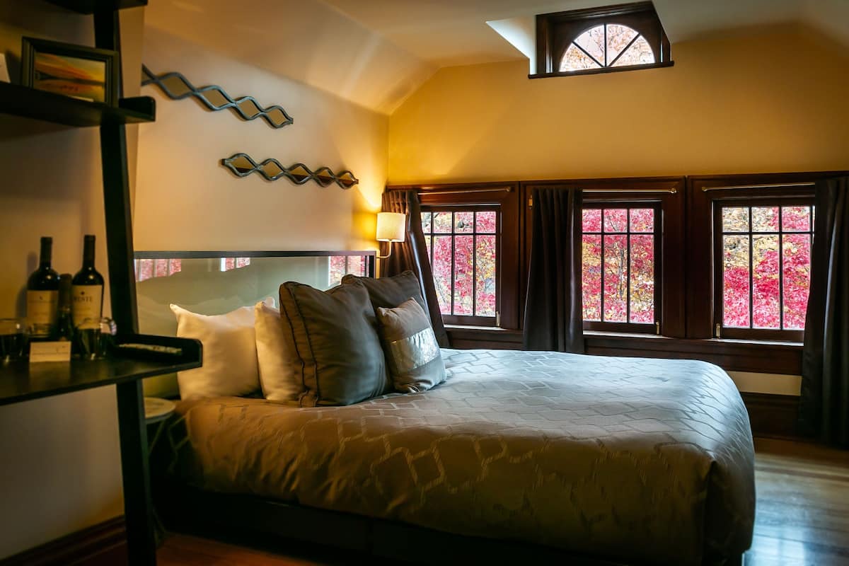A bedroom with a large bed, a nightstand with bottles, a wall clock, and windows with autumn foliage.