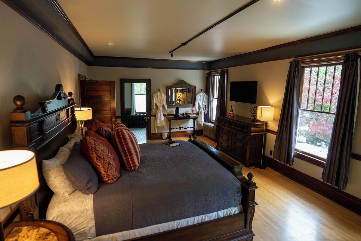 A spacious bedroom with a large bed, bedside lamps, a dresser, a vanity with a mirror, and a television.