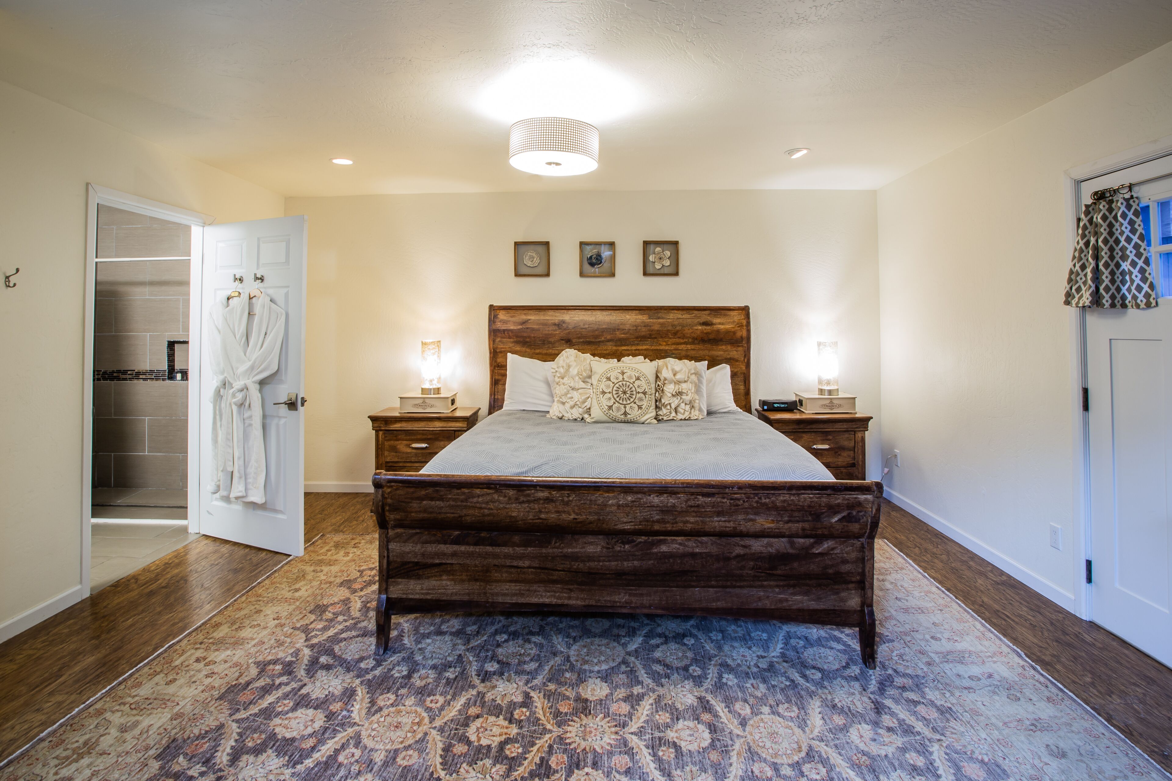 A bedroom with a wooden bed, a nightstand, a bedside table, a patterned rug, a closet with a robe hanging, and a ceiling light fixture.