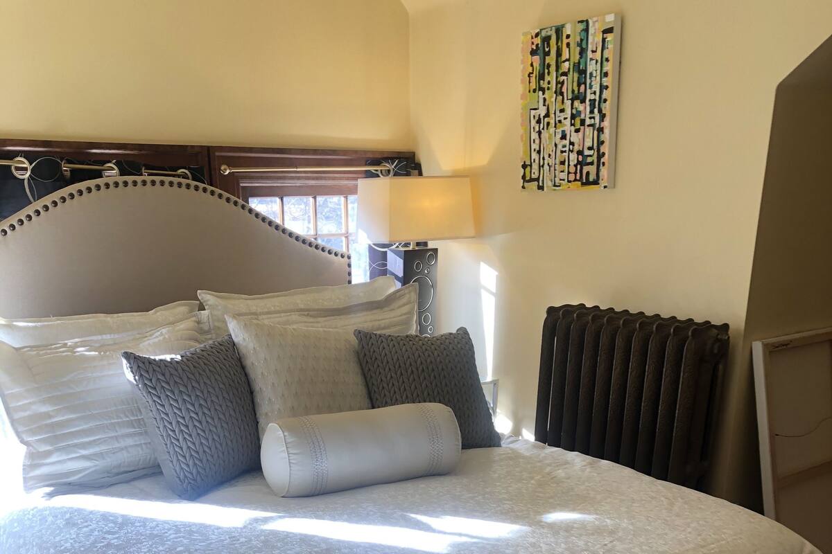 A bedroom with a large bed, multiple pillows, a bedside lamp, a radiator, and a window with curtains.