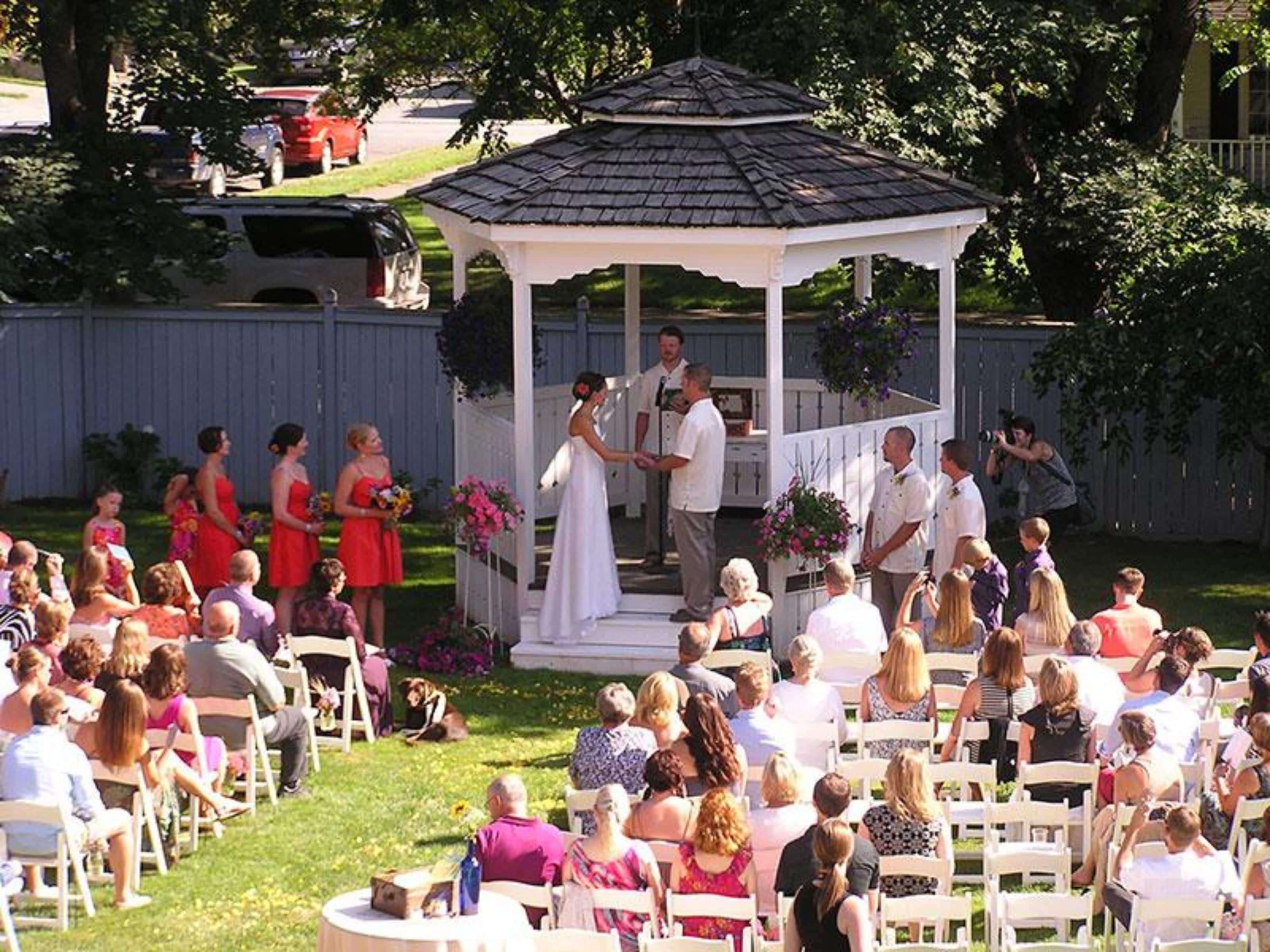 A gazebo wedding venue with guests seated on white chairs, a white fence, and a lawn.