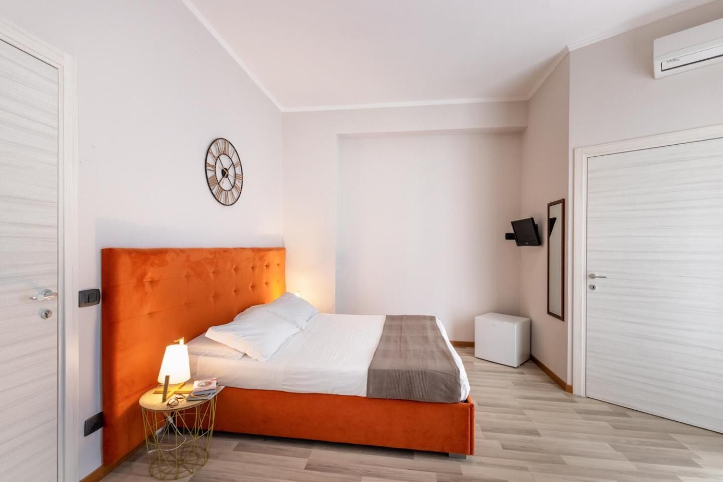 Een slaapkamer met een groot bed, een nachtkastje met lamp, een klok aan de muur en een klein wit kastje.